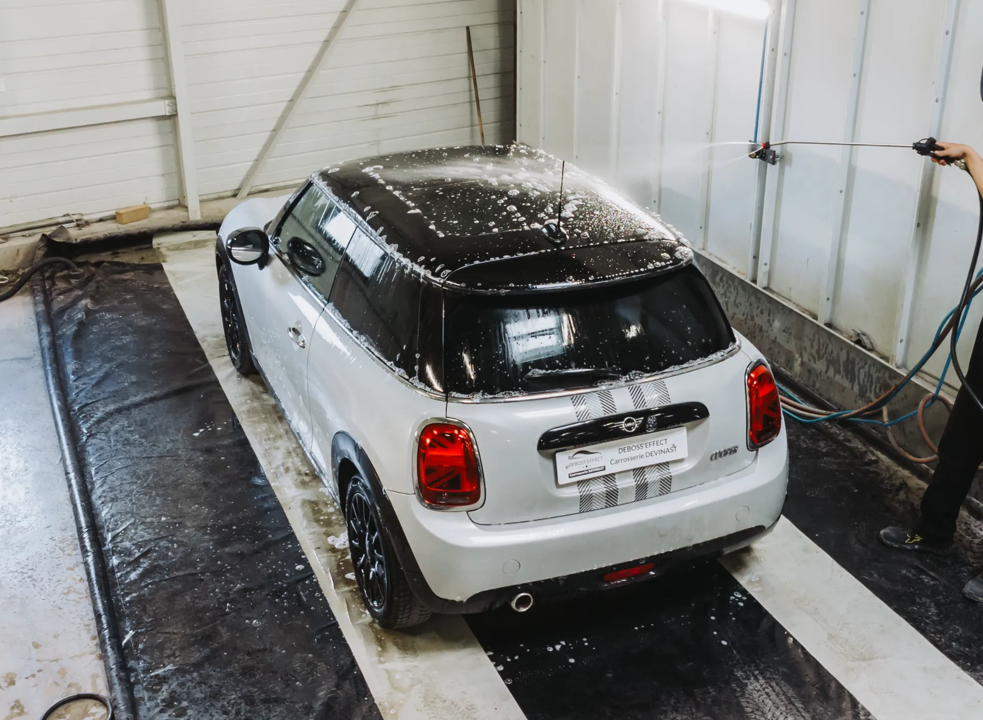 Une Mini Cooper blanche est lavée à l'intérieur de la carrosserie Devinast à Brognard. De la mousse recouvre le toit et la carrosserie, tandis qu'un jet d'eau est appliqué sur le toit.